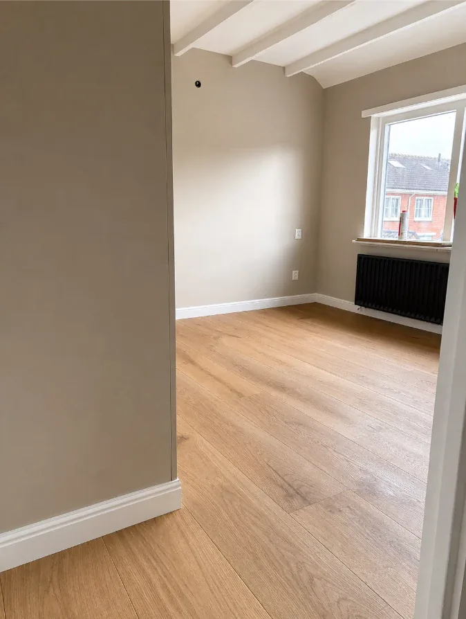 Kamer met balkenplafond en eiken parketvloer, met de raampartij en radiator aan de rechterzijde
